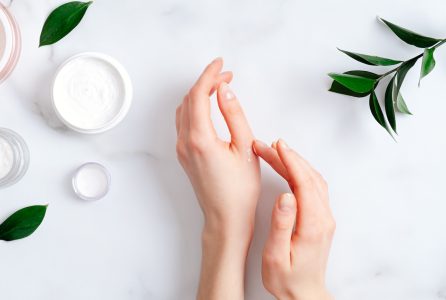 Cosmetic cream on female hands, jars with milk swirl cream and green leaves on white marble table. Flat lay, top view. Woman applying organic moisturizing hand cream. Hand skin care concept